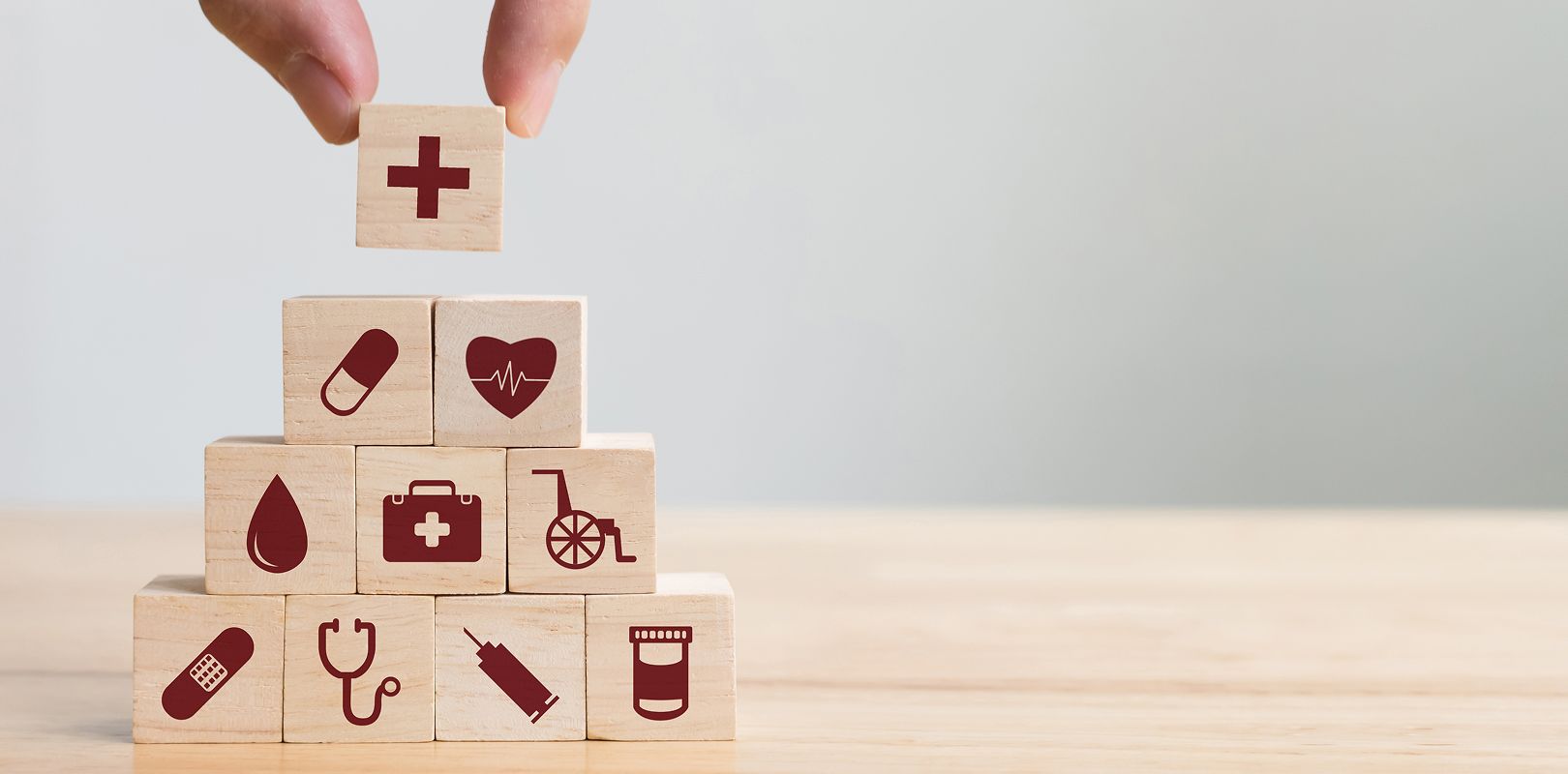 Eine Hand setzt einen roten Kreuz-Baustein auf eine Pyramide aus Holzklötzen mit Gesundheits-Symbolen wie Stethoskop und Rollstuhl.